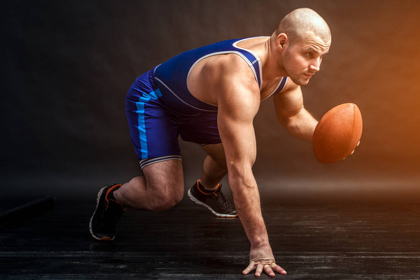 Um jovem atlético de meia-calça de luta azul e calções azuis está em uma posição baixa e segura uma bola de rugby marrom em um fundo preto isolado em um estúdio de fotos
 - Foto, Imagem