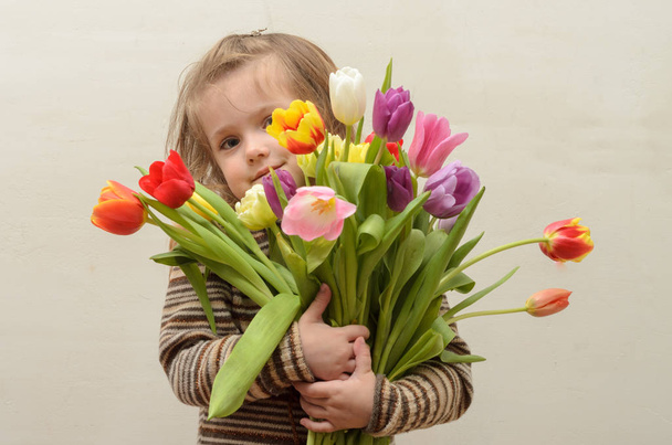 Happy baby girl rejoices and smiles with a bouquet of multi-colored tulips in hands - Фото, изображение