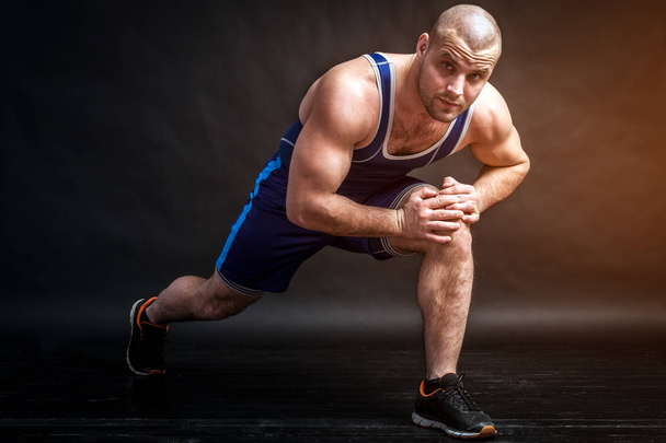 Een atletische jongeman in blauw worstelen panty's en blauwe shorts staat in een lage positie, maakt aanvallen en stretching op een zwarte geïsoleerde achtergrond in een fotostudio - Foto, afbeelding