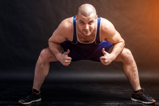A young athletic man in blue wrestling tights and blue shorts makes a squat with a wide leg arrangement and shows thumbs up on a black isolated background in a photo studio - Zdjęcie, obraz