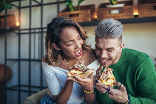 Змішана пара гонок їсть піцу в сучасному кафе. Вони сміються і їдять піцу і чудово проводять час
. - Фото, зображення