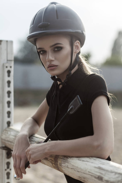 portrait of jockey with perfect skin and makeup wearing helmet leaning on obstacle in hippodrome, sport and fashion concept   - Fotó, kép