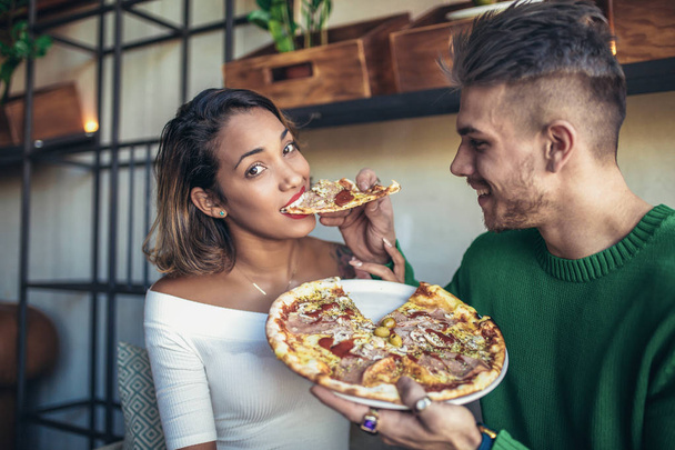 Para mieszanej rasy jedzenie pizzy w nowoczesną kawiarnię. Są one śmiejąc się i jedzenie pizzy i świetnie się bawimy. - Zdjęcie, obraz