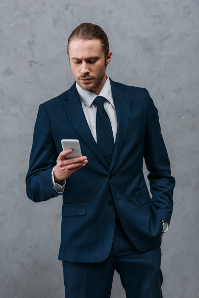 handsome confident businessman using smartphone in front of concrete wall - Zdjęcie, obraz