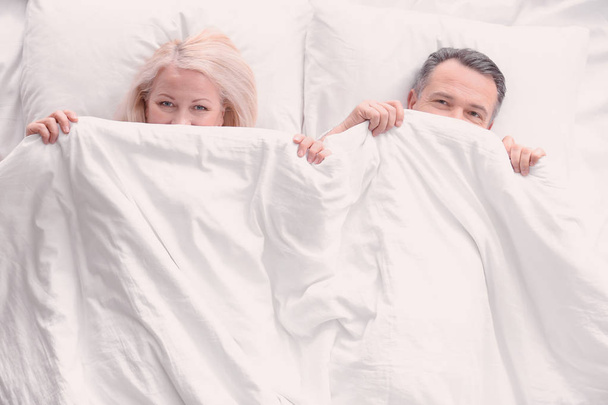 Senior couple hiding under blanket in bed together, top view - 写真・画像