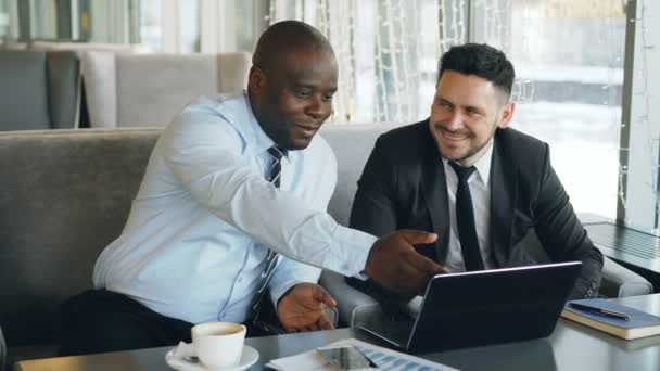 Cheerful African American businessman in formal clothes discussing business project with his caucasian colleague on his laptop in cafe - Filmmaterial, Video