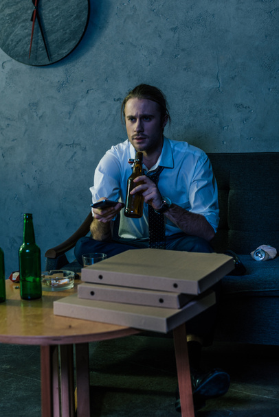 νέοι αλκοόλ εθισμένος άνθρωπος σε λευκό πουκάμισο, βλέποντας τηλεόραση και πίνοντας μπύρα μετά τη δουλειά στο καναπέ - Φωτογραφία, εικόνα