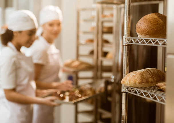panaderos que trabajan en la fabricación de hornear con panes frescos acostados en los estantes en primer plano
 - Foto, Imagen