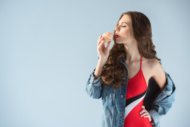 Πλάγια όψη της σαγηνευτική γυναίκα με κόκκινο μαγιό τρώει cupcake που απομονώνονται σε γκρι - Φωτογραφία, εικόνα