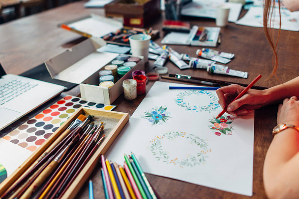 Közeli kép a női designer rajz virág kompozíciók a ceruzák ül körül a festék, gouache, ecsetek, és más művészeti munkahelyen kellékek - Fotó, kép