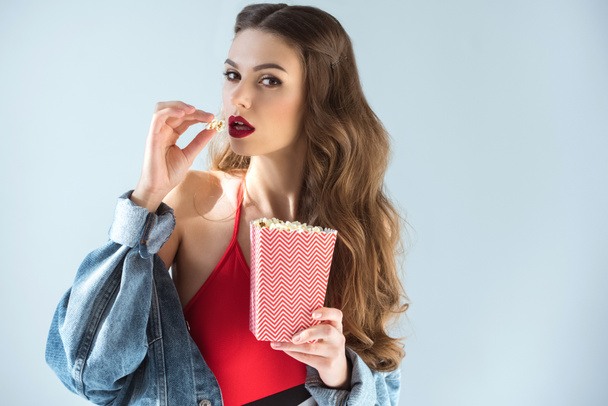 σαγηνευτικό κορίτσι με κόκκινα χείλη τρώγοντας ποπ κορν και βλέπουν τα φωτογραφικών μηχανών που απομονώνονται σε γκρι - Φωτογραφία, εικόνα