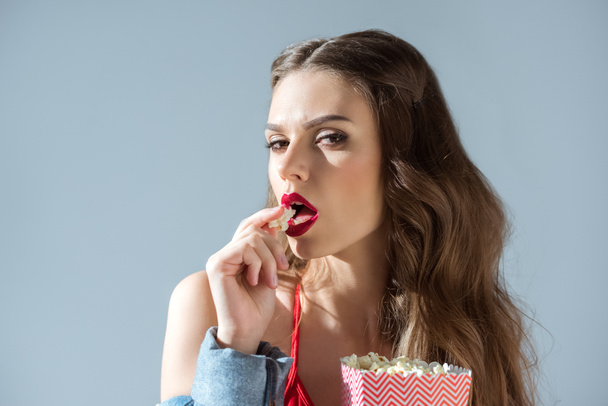 соблазнительная девушка ест попкорн и смотрит на камеру, изолированную от серого
 - Фото, изображение