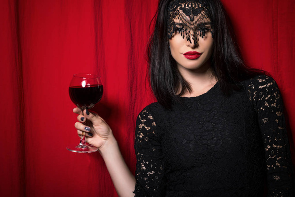 Young beautiful woman holding glass of wine over red curtains - Foto, afbeelding
