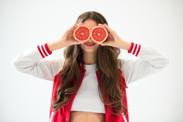 sexy girl in baseball jacket covering eyes with grapefruit pieces isolated on white - Photo, Image