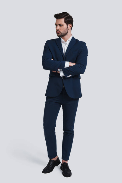full length of handsome young man in suit looking away and keeping arms crossed in studio - Foto, afbeelding