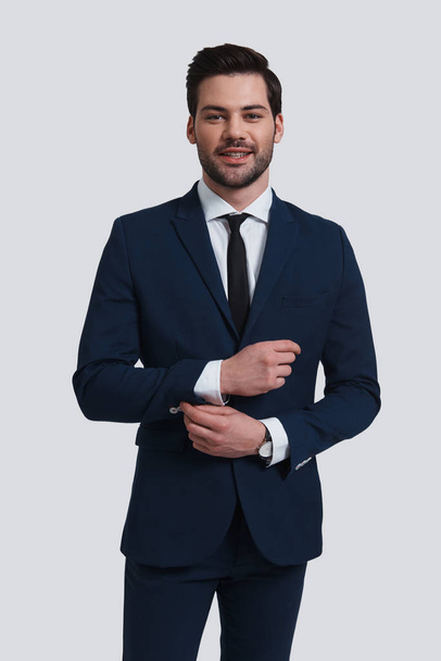 young man in suit adjusting sleeve and looking at camera with smile while standing against grey background - Photo, image