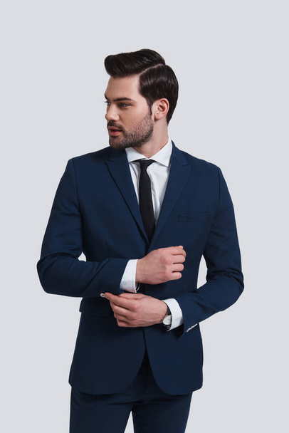 handsome young man in suit adjusting sleeve and looking away while standing against grey background - Φωτογραφία, εικόνα