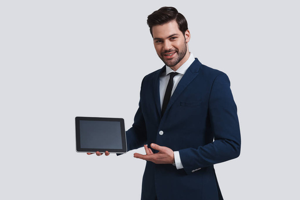 smiling man in full suit pointing at digital tablet and looking at camera with smile while standing against grey background - Foto, Imagem
