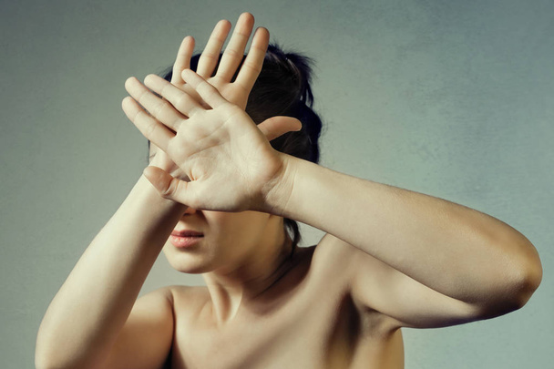 Concept of fear, shame, domestic violence. Woman covers her face by hands on light background. - Фото, изображение