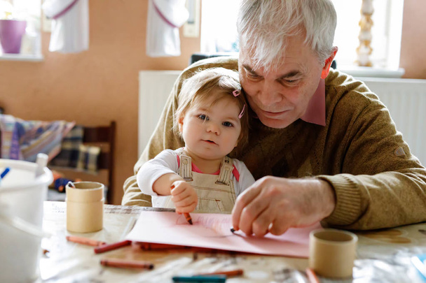 かわいい赤ちゃんの幼児の女の子と自宅でカラフルな鉛筆でハンサムなシニア祖父の絵画。孫と男は一緒に楽しんでる - 写真・画像
