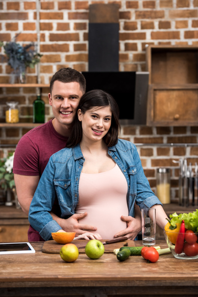 feliz joven embarazada pareja abrazando y sonriendo a la cámara mientras cocinan juntos
  - Foto, Imagen