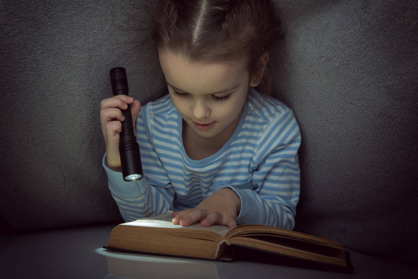 Pieni tyttö lukee satuja kirjan peiton alla aattona
 - Valokuva, kuva