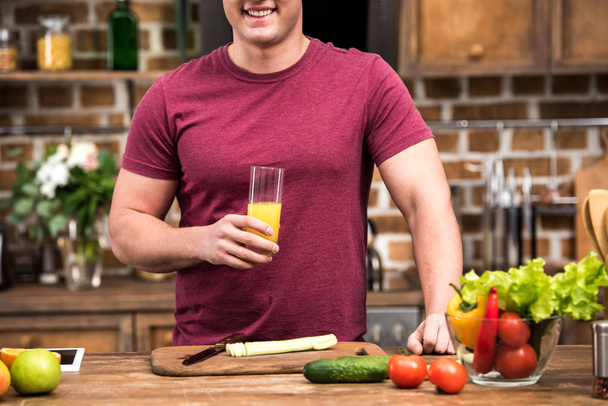 cropped shot of smiling young man holding glass of fresh orange juice at kitchen - Foto, afbeelding