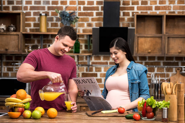 smiling young man pouring orange juice and happy pregnant woman reading travel newspaper - Photo, image
