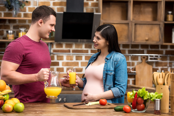 happy young pregnant couple holding glass and jug of fresh orange juice and smiling each other at kitchen - Photo, Image