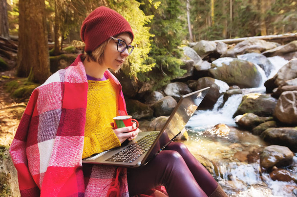 Hipster girl designer with a plaid on her shoulders in a hat and a yellow sweater and glasses with a laptop on her lap and a mug of coffee in her hands, look at the laptop screen sitting on a stone in - Fotó, kép