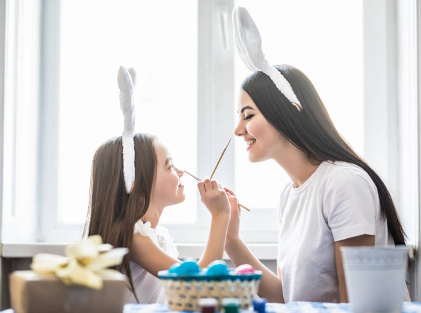 Vonzó fiatal nő, aranyos kislány húsvéti ünnep készül. Anya és lánya visel nyuszi füle szórakoznak egymással festett húsvéti nyuszi - Fotó, kép