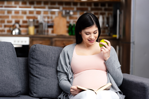 улыбающаяся молодая беременная женщина ест яблоко и читает дома книгу
 - Фото, изображение