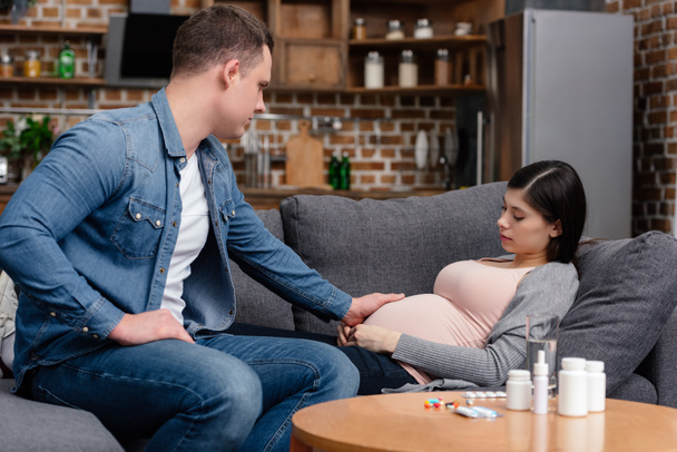 man touching belly of pregnant woman lying on sofa and various pills on table - Valokuva, kuva