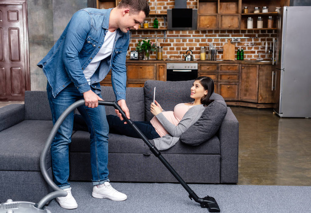young man cleaning carpet with vacuum cleaner while pregnant woman lying with digital tablet on sofa - Φωτογραφία, εικόνα