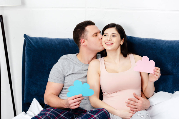 happy young pregnant couple holding blue and pink clouds symbols and kissing on bed - Фото, изображение