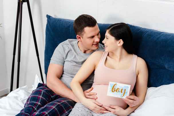 heureux jeune couple enceinte souriant sur le lit et tenant la carte avec mot garçon
 - Photo, image