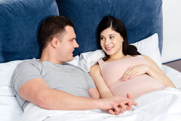 heureux jeune couple enceinte couché ensemble dans le lit et se regardant
 - Photo, image