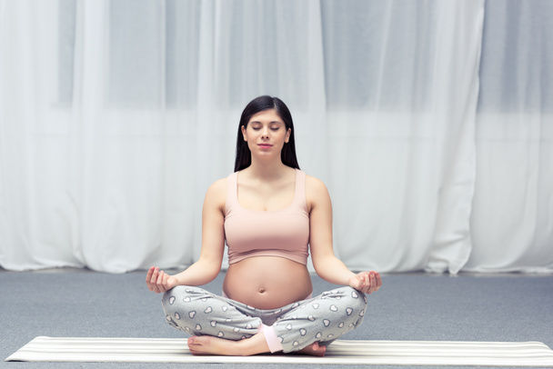 young pregnant woman with closed eyes sitting in lotus position on yoga mat - Photo, image