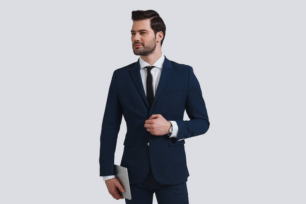 good looking young man in full suit adjusting his jacket and looking away with smile while standing against grey background - Foto, imagen