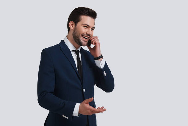 happy good looking young businessman in full suit talking on his smart phone while standing against grey background - Φωτογραφία, εικόνα