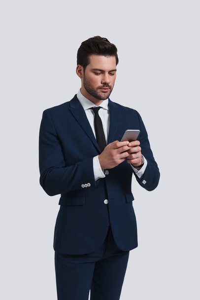 good looking young man using smart phone while standing against grey background - Фото, изображение