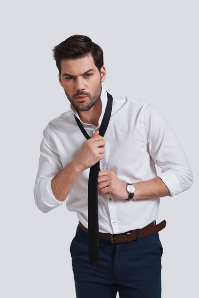 tired young man looking at camera and taking on his necktie while standing against grey background - Zdjęcie, obraz