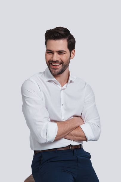 cheerful and handsome man keeping arms crossed and looking away while sitting against grey background - Foto, afbeelding