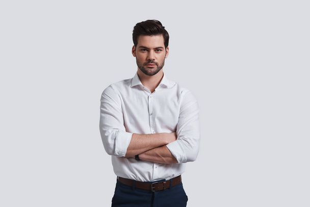 young man looking at camera and keeping arms crossed while standing against grey background - Foto, afbeelding