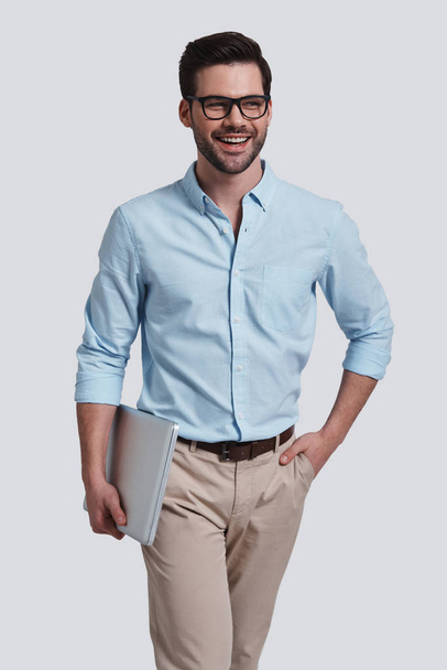 smiling businessman keeping hand in pocket and holding laptop while posing in studio - Fotoğraf, Görsel