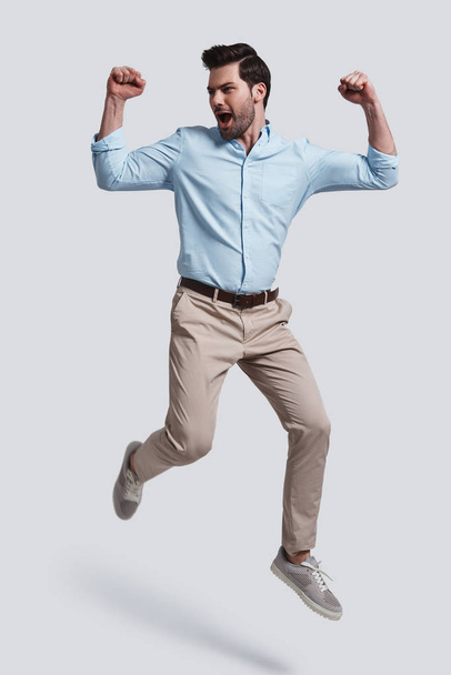 full length of handsome young man gesturing success and shouting while jumping grey background - Fotoğraf, Görsel