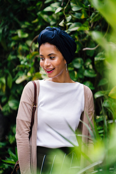 Portrait head shot of a beautiful, young, elegant Muslim woman in a stylish blue turban headscarf (hijab). She is smiling against a blurred green background of plants in the day. - Photo, image