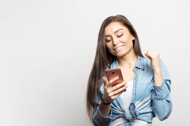 Portrait of young smiling woman with pink smartphone receiving some great news over white background. Lifestyle, people and technology concept. - 写真・画像