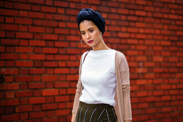 Portrait of a stylish Muslim fashionista influencer blogger woman. She is elegant, beautiful and wearing a blue turban as she stands against a red brick wall in the day. She is young and beautiful. - Fotoğraf, Görsel