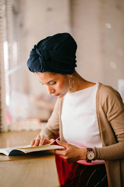 young, attractive Muslim woman (Arab, Malay, Asian) reads a book by the window of a cafe in the day. She is wearing a turban (headscarf, hijabi) and is elegantly dressed in earthy tones. - 写真・画像